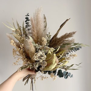 Puede incluir: Un ramo de flores secas con una variedad de texturas y colores. El arreglo incluye hierba de la pampa, eucalipto y otras flores secas en tonos beige, verde y marr&oacute;n. El ramo se sostiene en la mano sobre un fondo liso.