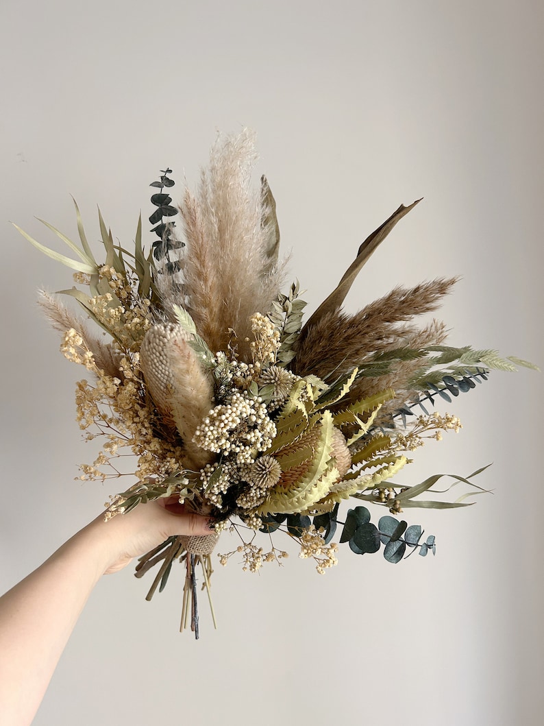 Puede incluir: Un ramo de flores secas con una variedad de texturas y colores. El arreglo incluye hierba de la pampa, eucalipto y otras flores secas en tonos beige, verde y marr&oacute;n. El ramo se sostiene en la mano sobre un fondo liso.