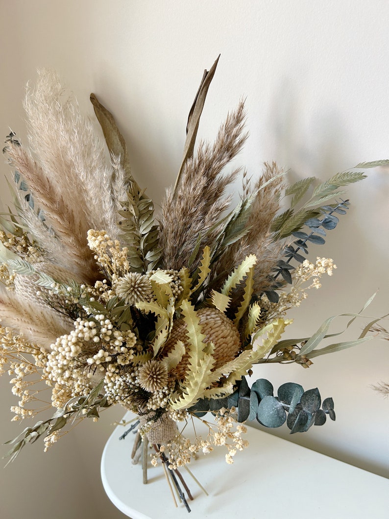 Puede incluir: Un ramo de flores secas con hierba de la Pampa, eucalipto y otras plantas secas en tonos beige, marr&oacute;n y verde. El ramo est&aacute; atado con hilo y arpillera, creando una est&eacute;tica natural y r&uacute;stica.