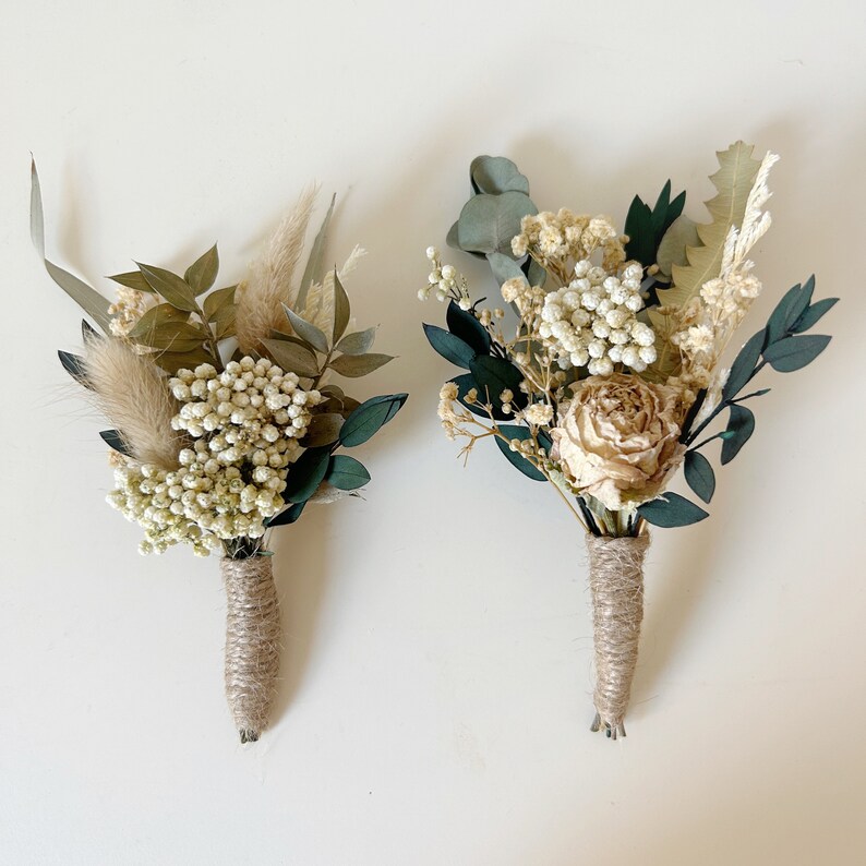 Puede incluir: Dos prendidos con flores y vegetaci&oacute;n secas. Uno presenta una rosa beige, el otro un elemento esponjoso similar al trigo. Ambos est&aacute;n envueltos con hilo de c&aacute;&ntilde;amo, creando una est&eacute;tica r&uacute;stica y natural. Perfectos para bodas o eventos especiales.
