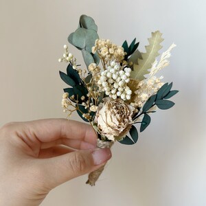 Puede incluir: Un peque&ntilde;o prendedor floral seco con una rosa color crema, paniculata blanca y hojas de eucalipto verde oscuro. El arreglo est&aacute; atado con hilo de bramante e incluye acentos de follaje seco. Perfecto para bodas o eventos especiales.