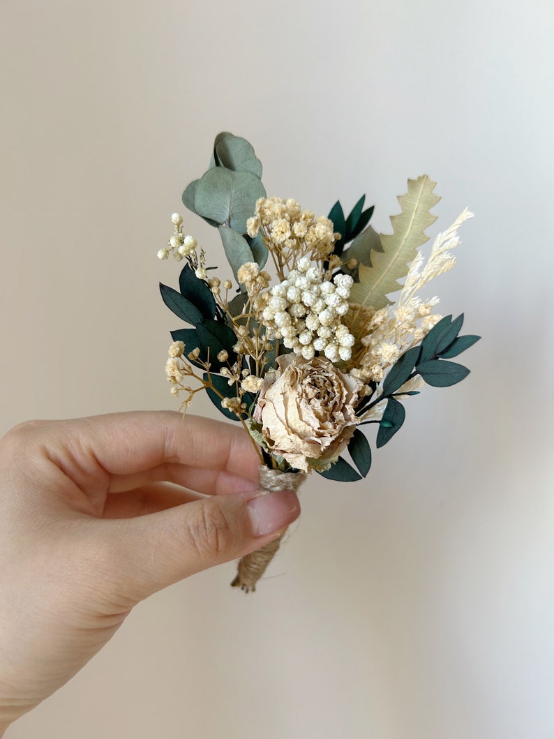 Puede incluir: Un peque&ntilde;o prendedor floral seco con una rosa color crema, paniculata blanca y hojas de eucalipto verde oscuro. El arreglo est&aacute; atado con hilo de bramante e incluye acentos de follaje seco. Perfecto para bodas o eventos especiales.