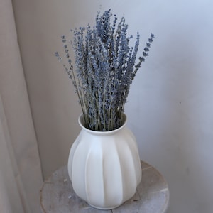 May include: A white, ribbed ceramic vase filled with dried lavender. The lavender has slender stems and small, blue-purple flower heads. The vase sits on a light-colored wooden surface, creating a simple, natural aesthetic.