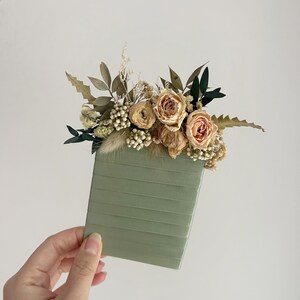 Puede incluir: Un cuaderno verde salvia adornado con flores secas. El arreglo floral incluye rosas color melocot&oacute;n, bayas blancas y varios follajes verdes y beige. El cuaderno tiene una textura acanalada.