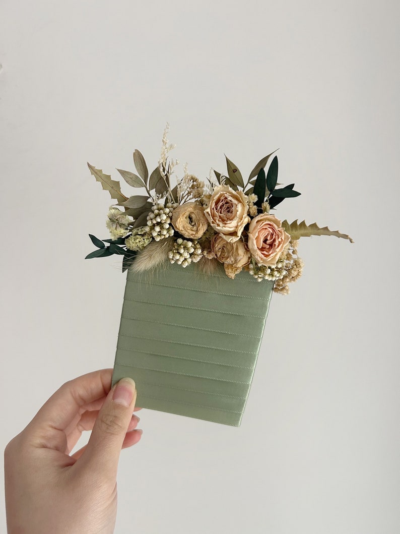 Puede incluir: Un cuaderno verde salvia adornado con flores secas. El arreglo floral incluye rosas color melocot&oacute;n, bayas blancas y varios follajes verdes y beige. El cuaderno tiene una textura acanalada.