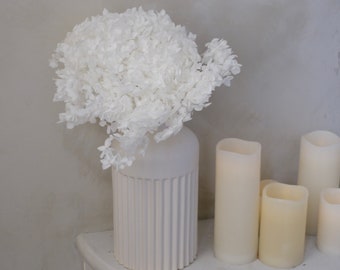 May include: A white ceramic vase with vertical ridges holds a large arrangement of white flowers. Several ivory-colored pillar candles are arranged to the right of the vase, creating a minimalist, elegant display.