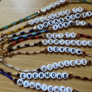 Può includere: Una collezione di braccialetti di perline con perline bianche che compongono i nomi degli album di Taylor Swift, tra cui "Debut", "Fearless", "Speak Now", "1989", "Red", "Reputation", "Lover", "Folklore", "Evermore" e "Midnights".