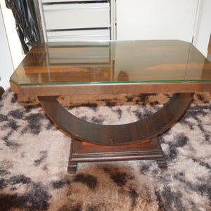May include: A rectangular side table featuring a dark brown wooden frame with a smooth glass top. The base consists of a large, curved, crescent-shaped wooden support that connects the tabletop to a flat, rectangular wooden pedestal base with feet.