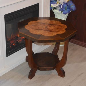 May include: A hexagonal wooden side table featuring a dark brown frame and a lighter, swirling burl wood inlay on the tabletop. The table is supported by four curved legs and includes a smaller hexagonal shelf positioned between the legs near the base.