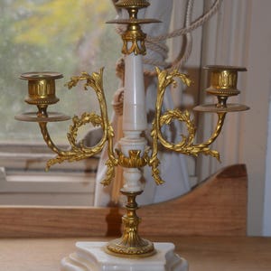 May include: A three-light candelabra featuring a white stone base and central column with ornate gold-toned metal arms and candle cups. The base is supported by four small gold-toned feet, and the metal components display intricate leaf-like detailing.