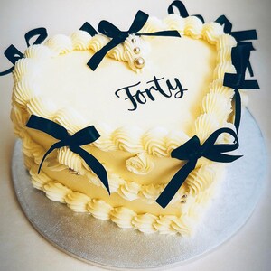 May include: Heart-shaped cake with buttercream frosting and black bows. The cake is decorated with the word "Forty" in black script and gold and silver sprinkles. The cake sits on a silver cake board.