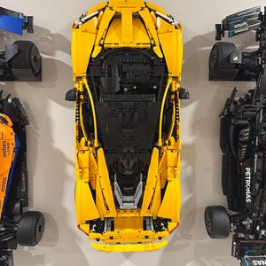 May include: A top-down view of a yellow Lego race car model, with black undercarriage details. The car is flanked by other Lego race car models in blue, black, and orange. The models are mounted on a light gray wall.