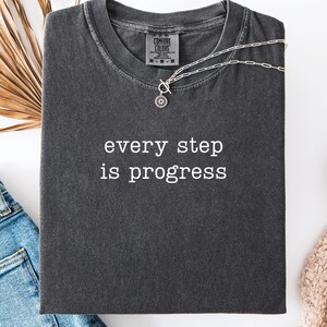 May include: A dark gray t-shirt with the phrase "every step is progress" in white lettering. A silver necklace with a sun pendant rests on the shirt. The word "Pepper" is at the bottom left corner of the image.