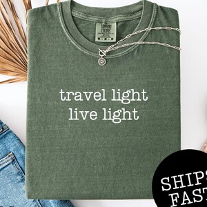 May include: A moss green t-shirt with the words "travel light live light" in white. A silver necklace with a pendant is on the shirt. The shirt is folded with a pair of blue jeans and a white shoe visible.