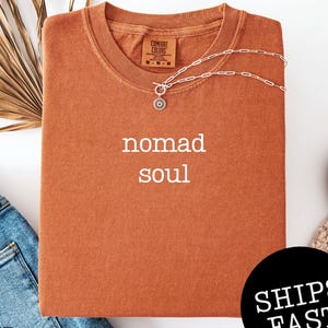 May include: A rust-colored t-shirt with the words "nomad soul" printed in white. A silver necklace with a circular pendant is draped across the shirt. The shirt is folded and placed on a white surface next to a pair of blue jeans.