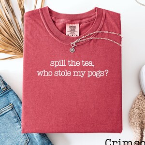 May include: Crimson-colored t-shirt with the text "spill the tea, who stole my pogs?" in white. A silver necklace with a circular pendant rests on the shirt. The shirt is folded next to a pair of blue jeans and a white shoe.