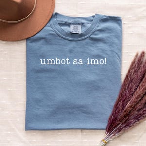 May include: A dusty blue t-shirt with the text "umbot sa imo!" in white. A brown hat and a bundle of dried, reddish-purple pampas grass are also in the image. The shirt is a Comfort Colors brand.