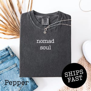 May include: A dark gray t-shirt with the words "nomad soul" in white. A silver necklace with a sun pendant rests on the shirt. The shirt is folded on top of blue jeans. A black circle with the words "SHIPS FAST" is visible.