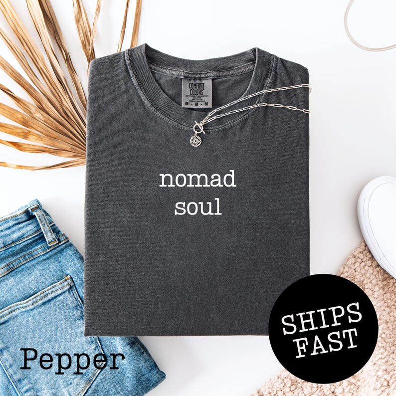 May include: A dark gray t-shirt with the words "nomad soul" in white. A silver necklace with a sun pendant rests on the shirt. The shirt is folded on top of blue jeans. A black circle with the words "SHIPS FAST" is visible.