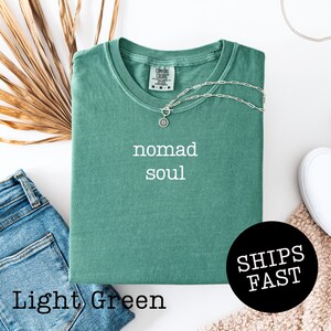 May include: Light green t-shirt with the words "nomad soul" in white. A silver necklace with a pendant rests on the shirt. The shirt is folded, with a pair of blue jeans and a white shoe visible. The words "SHIPS FAST" are in a black circle.