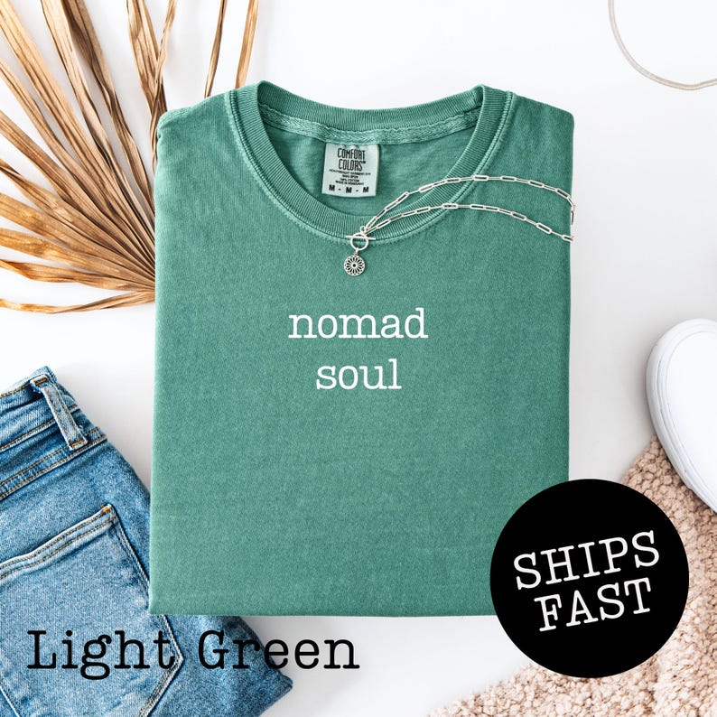 May include: Light green t-shirt with the words "nomad soul" in white. A silver necklace with a pendant rests on the shirt. The shirt is folded, with a pair of blue jeans and a white shoe visible. The words "SHIPS FAST" are in a black circle.