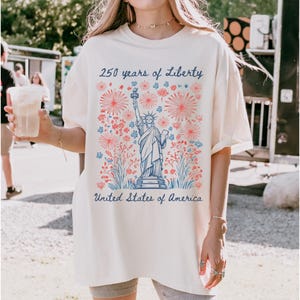 Peut inclure: T-shirt cr&egrave;me avec un motif de la Statue de la Libert&eacute;, des motifs floraux et de feux d'artifice rouges et bleus. Le texte indique "250 years of Liberty" et "United States of America" en &eacute;criture bleue.