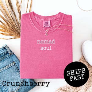 May include: A Crunchberry-colored t-shirt with the words "nomad soul" in white. A silver necklace with a pendant is on the shirt. The shirt is folded with a pair of blue jeans and a white shoe visible.