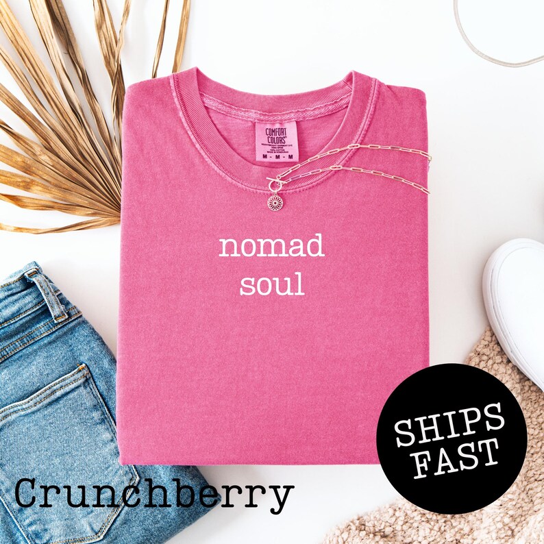 May include: A Crunchberry-colored t-shirt with the words "nomad soul" in white. A silver necklace with a pendant is on the shirt. The shirt is folded with a pair of blue jeans and a white shoe visible.