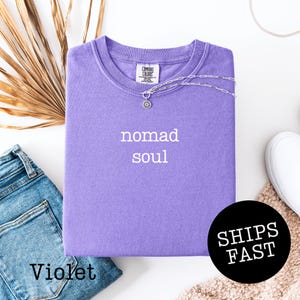 May include: A violet Comfort Colors t-shirt with the words "nomad soul" printed in white. A silver necklace with a pendant is draped across the shirt. The word "Violet" is printed at the bottom left.