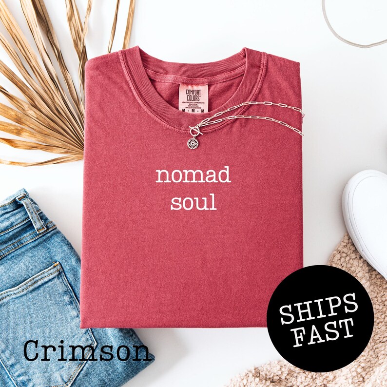 May include: Crimson-colored t-shirt with the words "nomad soul" in white. A silver necklace with a pendant rests on the shirt. The shirt is folded, with a pair of blue jeans and a white shoe visible. A black circle with "SHIPS FAST" is in the image.