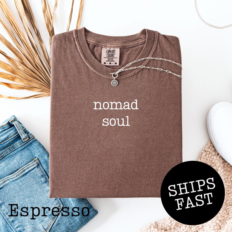 May include: Espresso-colored t-shirt with the words "nomad soul" in white. A silver necklace with a sun pendant rests on the shirt. The shirt is folded, with a pair of blue jeans and a "Ships Fast" sticker nearby.