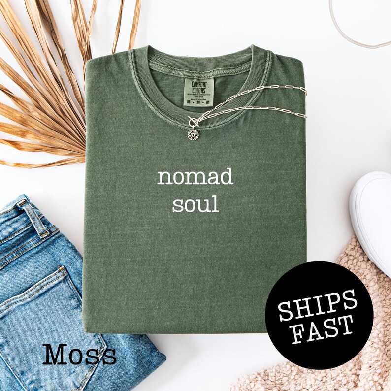May include: A moss green t-shirt with the words "nomad soul" in white. A silver necklace with a pendant is on the shirt. The shirt is folded next to a pair of blue jeans. A black circle with the words "SHIPS FAST" is in the image.