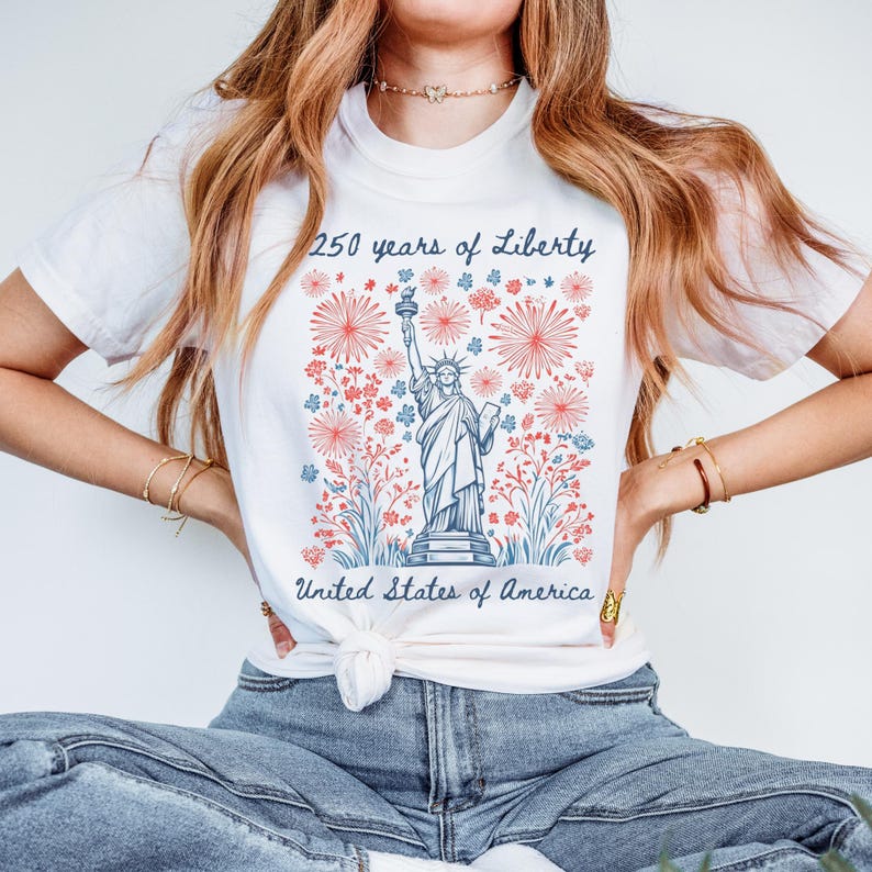 May include: White t-shirt featuring a graphic of the Statue of Liberty with the text "250 years of Liberty" and "United States of America". The design includes red and blue floral and firework elements.