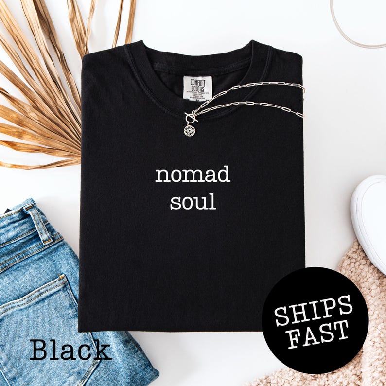 May include: Black t-shirt with the words "nomad soul" in white. A silver necklace with a pendant is draped on the shirt. The words "SHIPS FAST" are in a black circle. The word "Black" is at the bottom left.