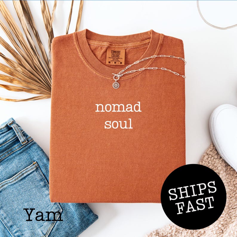 May include: A rust-colored t-shirt with the words "nomad soul" printed in white. A silver necklace with a circular pendant is draped across the shirt. The shirt is folded and placed on a white surface next to a pair of blue jeans.