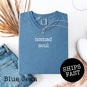 May include: A blue t-shirt with the words "nomad soul" in white. The shirt is folded and has a silver necklace draped over it. A pair of blue jeans and a white shoe are also in the image. The shirt has the text "SHIPS FAST" on a black circle.