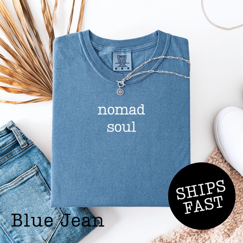 May include: A blue t-shirt with the words "nomad soul" in white. The shirt is folded and has a silver necklace draped over it. A pair of blue jeans and a white shoe are also in the image. The shirt has the text "SHIPS FAST" on a black circle.
