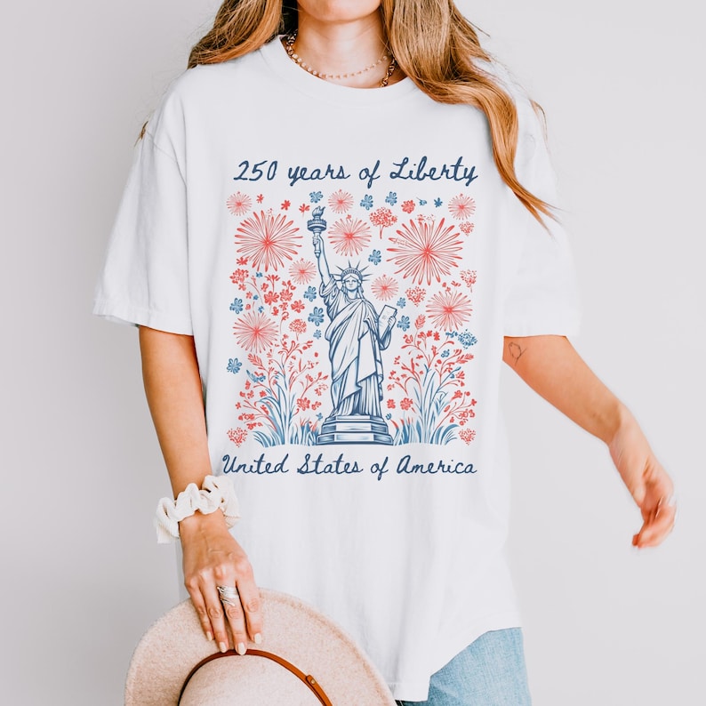 Peut inclure: T-shirt blanc avec un motif de la Statue de la Libert&eacute;, entour&eacute;e de motifs floraux rouges et bleus. Le texte indique "250 years of Liberty" et "United States of America" en bleu. Le t-shirt a des manches courtes et une coupe ample.
