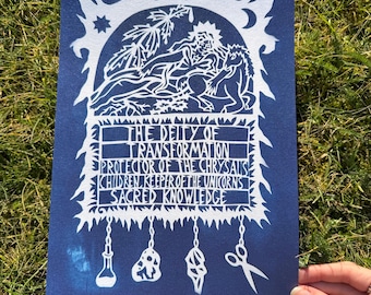 May include: A blue print with white ink depicting a deity of transformation, protector of the chrysalis, children keeper of the unicorns, sacred knowledge. The print features a stylized image of a person with a wolf, a crescent moon, a star, and a palm tree. There are five white objects hanging from the bottom of the print: a bottle, a rock, a crystal, a shell, and a pair of scissors.