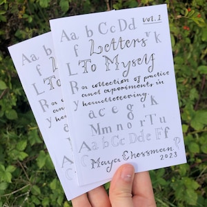 以下が含まれることがあります： 「Letters To Myself, Vol. 1」というタイトルの白い冊子と、手書きの文字と単語のコレクション。テキストには「a collection of practice and experiments in hand lettering.」と「Maya Chessman, 2023」も含まれています。