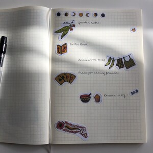 Puede incluir: Una p&aacute;gina de un planificador o diario con notas escritas a mano y pegatinas. Las notas incluyen "Garden Notes", "Books Read", "Housework to Do", "Plans for seeing friends" y "Recipes to try". Las pegatinas incluyen una vaina de guisantes, un libro, calcetines colgados en una cuerda para tender la ropa, cartas de juego, un taz&oacute;n de sopa, un cupcake y una pluma.