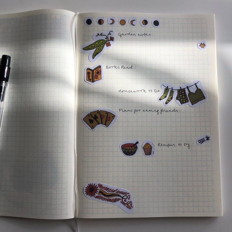 Puede incluir: Una p&aacute;gina de un planificador o diario con notas escritas a mano y pegatinas. Las notas incluyen "Garden Notes", "Books Read", "Housework to Do", "Plans for seeing friends" y "Recipes to try". Las pegatinas incluyen una vaina de guisantes, un libro, calcetines colgados en una cuerda para tender la ropa, cartas de juego, un taz&oacute;n de sopa, un cupcake y una pluma.