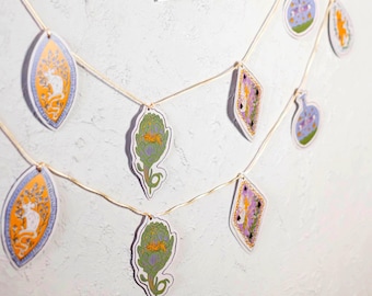 May include: A decorative garland featuring two rows of hanging paper cutouts suspended on a thin string. The cutouts include orange oval shapes with white mouse illustrations, green artichoke shapes, and diamond-shaped purple floral designs.