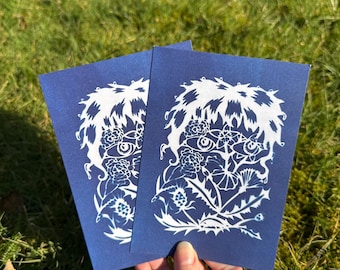 May include: Two blue cards with white ink illustrations of a stylized face surrounded by flowers and leaves. The face has a single eye and a nose.