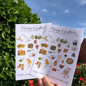 Puede incluir: Dos hojas de pegatinas con el texto "Home Comforts" y "journal/planner stickers" impreso en ellas. Las pegatinas presentan ilustraciones de varios objetos hogare&ntilde;os, incluyendo una tetera, una mecedora, una casa, una telara&ntilde;a, un rat&oacute;n, una bola de lana, una herradura, una magdalena, un libro, un coraz&oacute;n, un tr&eacute;bol, una abeja, una bota, una aguja de coser, un par de tijeras, un frasco, una olla, una taza de t&eacute;, un par de calcetines, una camisa a cuadros, un pantal&oacute;n, un frasco marr&oacute;n, una colcha y una baraja de cartas.