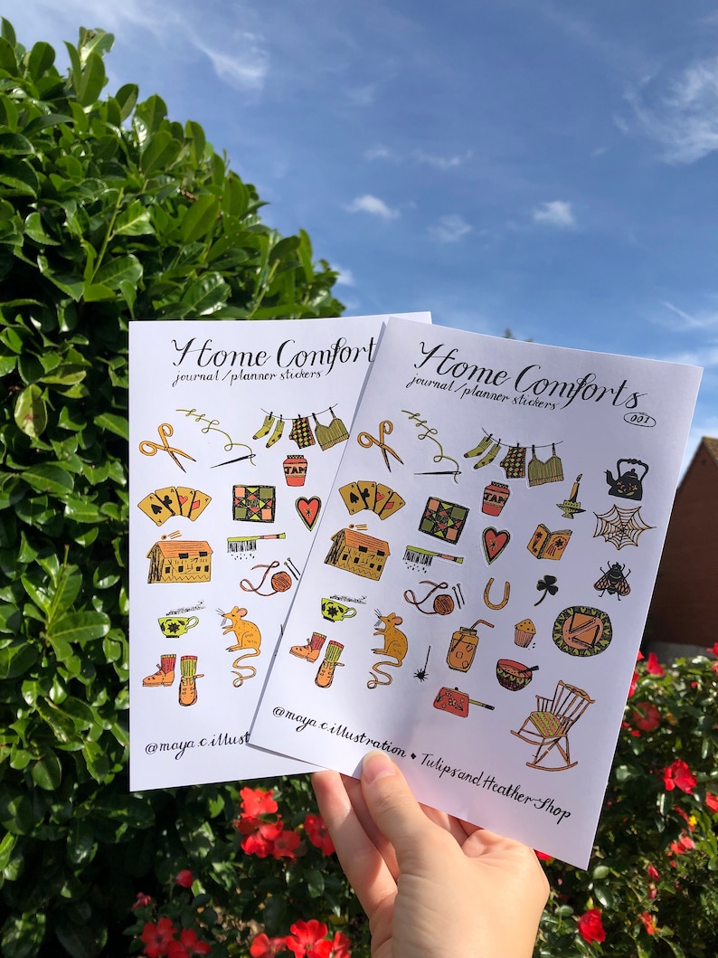 Puede incluir: Dos hojas de pegatinas con el texto "Home Comforts" y "journal/planner stickers" impreso en ellas. Las pegatinas presentan ilustraciones de varios objetos hogare&ntilde;os, incluyendo una tetera, una mecedora, una casa, una telara&ntilde;a, un rat&oacute;n, una bola de lana, una herradura, una magdalena, un libro, un coraz&oacute;n, un tr&eacute;bol, una abeja, una bota, una aguja de coser, un par de tijeras, un frasco, una olla, una taza de t&eacute;, un par de calcetines, una camisa a cuadros, un pantal&oacute;n, un frasco marr&oacute;n, una colcha y una baraja de cartas.