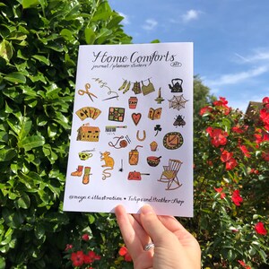 Puede incluir: Una hoja de pegatinas con ilustraciones de objetos hogare&ntilde;os acogedores, como una tetera, una mecedora, una casa, una telara&ntilde;a, un libro, una taza de t&eacute;, un tarro de mermelada, un coraz&oacute;n, una herradura, un rat&oacute;n, un par de botas, una vela, una aguja de coser, un carrete de hilo, un par de tijeras, un par de calcetines, una prenda de vestir colgada en una cuerda para tender la ropa, un tr&eacute;bol, una abeja, un muffin, un taz&oacute;n, una bola de lana, una llave, un jab&oacute;n y una taza. La hoja se titula "Home Comforts Journal/Planner Stickers 001" e incluye el nombre de usuario de las redes sociales "@maya.c.illustration" y "TulipsandHeatherShop".