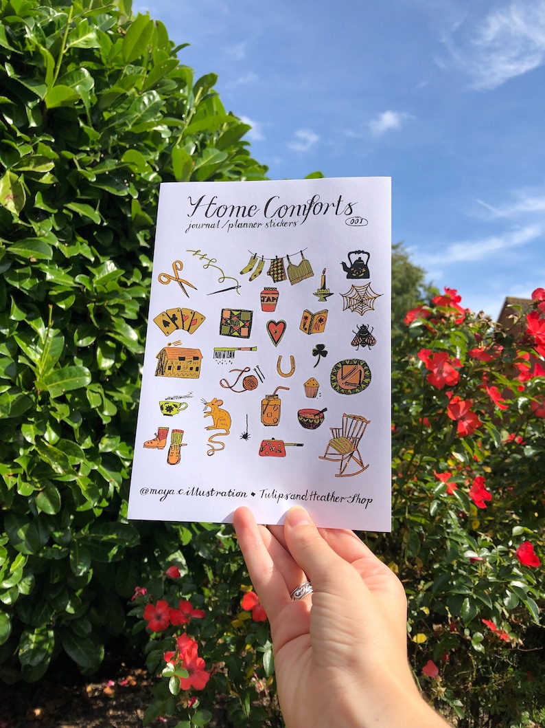 Puede incluir: Una hoja de pegatinas con ilustraciones de objetos hogare&ntilde;os acogedores, como una tetera, una mecedora, una casa, una telara&ntilde;a, un libro, una taza de t&eacute;, un tarro de mermelada, un coraz&oacute;n, una herradura, un rat&oacute;n, un par de botas, una vela, una aguja de coser, un carrete de hilo, un par de tijeras, un par de calcetines, una prenda de vestir colgada en una cuerda para tender la ropa, un tr&eacute;bol, una abeja, un muffin, un taz&oacute;n, una bola de lana, una llave, un jab&oacute;n y una taza. La hoja se titula "Home Comforts Journal/Planner Stickers 001" e incluye el nombre de usuario de las redes sociales "@maya.c.illustration" y "TulipsandHeatherShop".