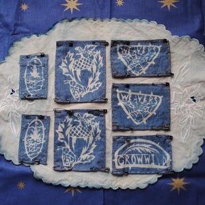 May include: Several denim patches with white designs, including a lighthouse, thistle, and text. The patches are pinned to a white, scalloped fabric on a blue background with gold stars. The patches vary in size and shape.