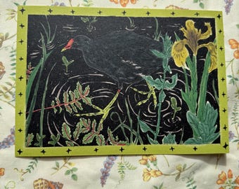 May include: A rectangular art print featuring a black bird with a red beak standing in water, surrounded by green plants and yellow flowers. The print has a green border with black star accents. The background is black.